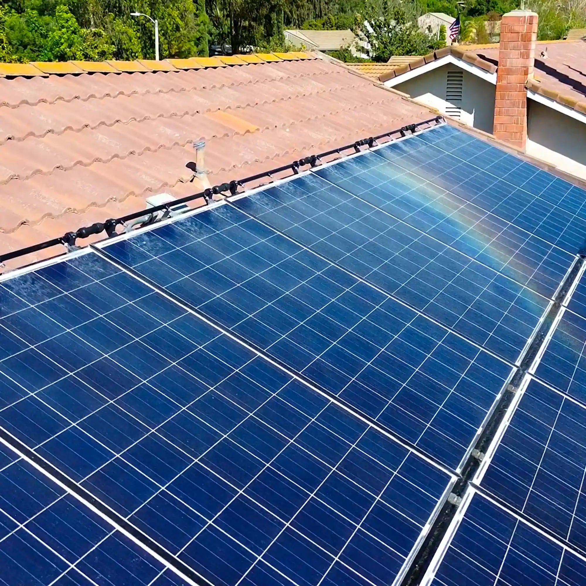 Solar panels on a residential roof being cleaned by the Home Elite Solar Panel Cleaning System with surrounding houses and greenery.